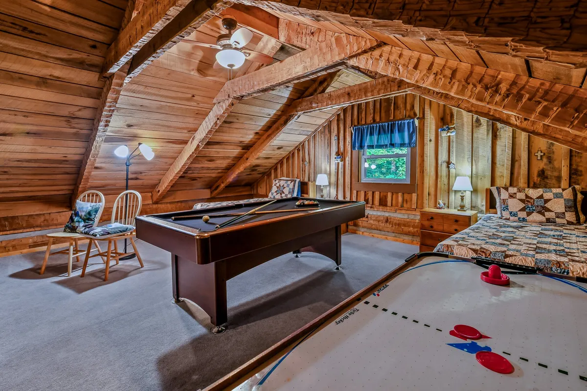 This image clearly displays a game room that features both a pool table and an air hockey table, as well as a bed, all beneath rustic wooden beams and with windows letting in natural light. The arrangement reflects an environment designed for fun, relaxation, and flexible accommodation.