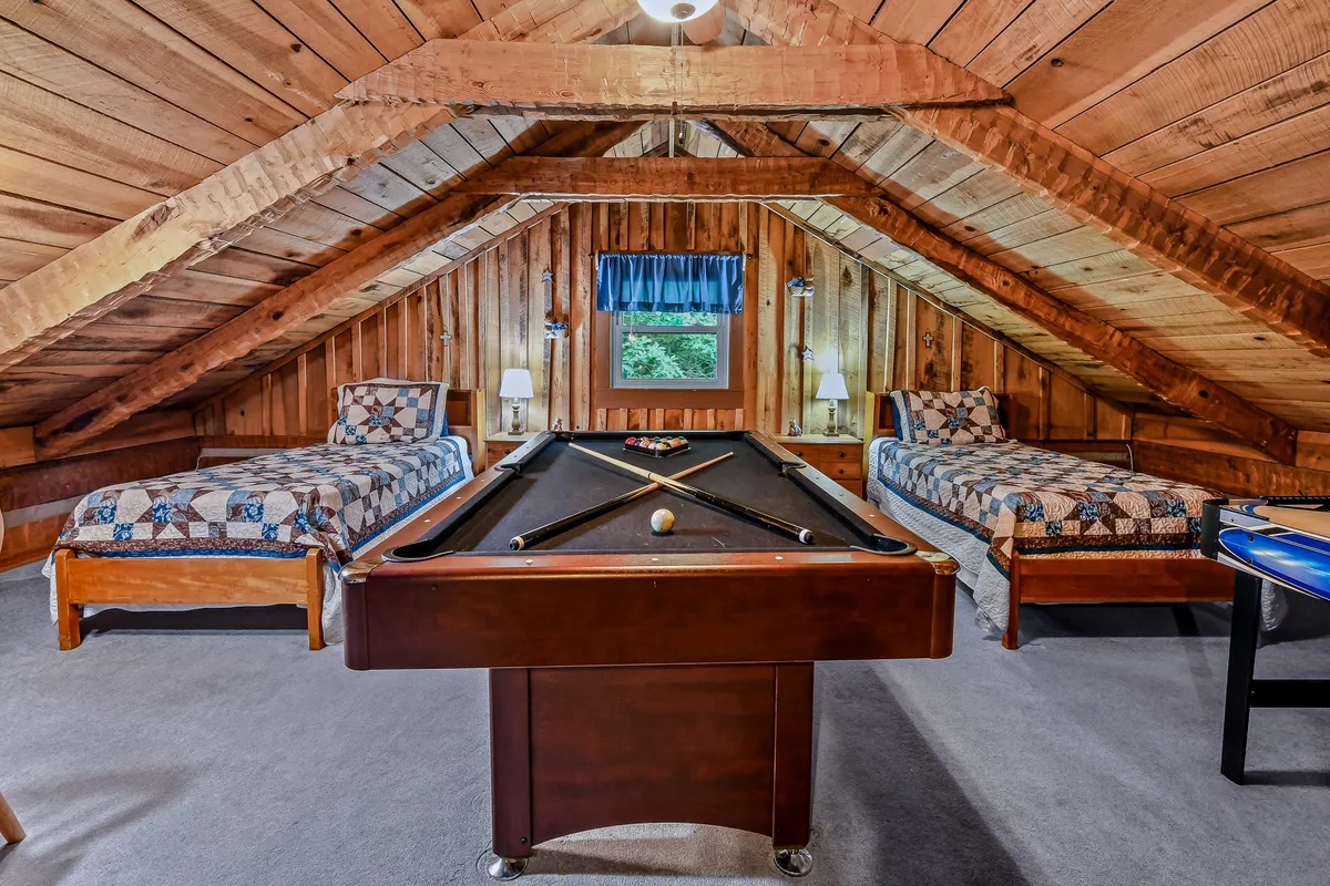 A playful, inviting room with a pool table at the center and a glimpse of bedrooms with quilts nearby, set under wooden beams. The scene suggests an open, multi-functional space perfect for both games and restful moments.