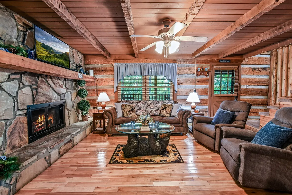 A cozy living area featuring a prominent stone fireplace, comfortable floral-patterned sofa, a cabin-style wooden ceiling, and a glass coffee table. The open layout hints at a space perfect for gathering, relaxing by the fire, and socializing after a day out exploring local attractions.