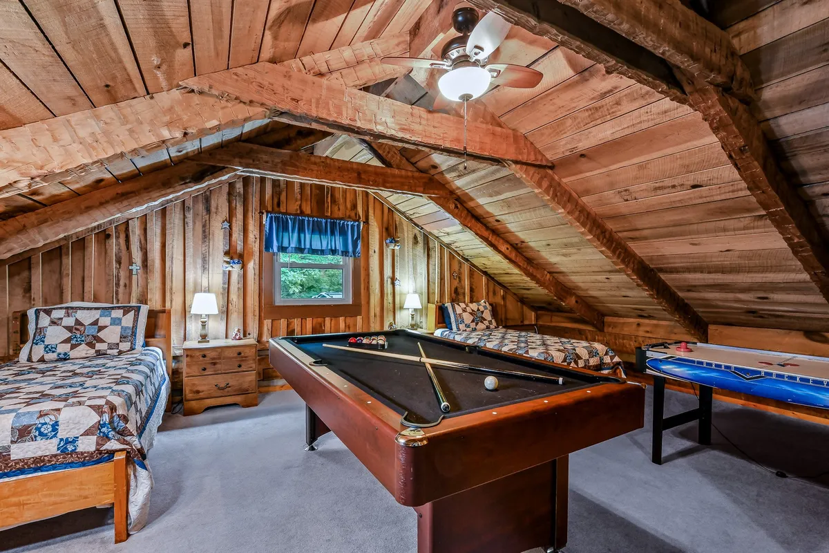 A multifunctional game room with a pool table and separate bedding area, accented by exposed wooden beams and a window that lets in natural light. The scene is playful and relaxed, highlighting versatile sleeping options and opportunities for leisure.