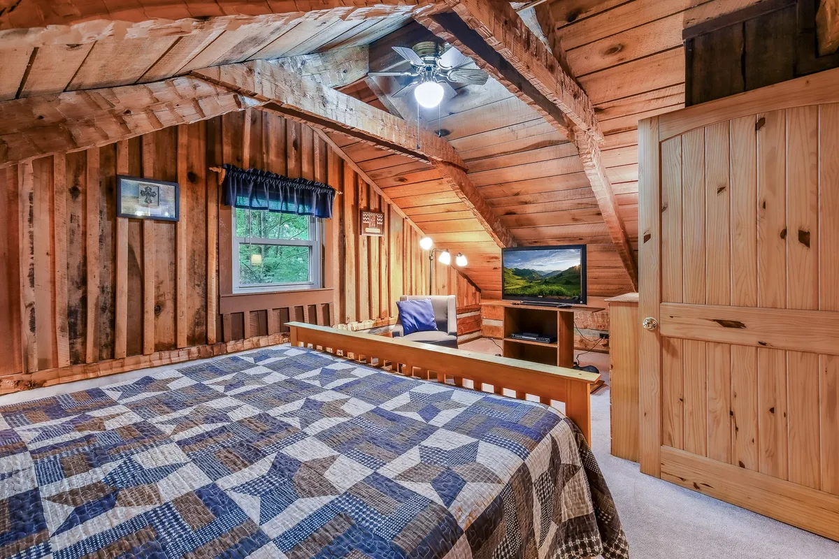 A comfortable bedroom with a wooden bed, plaid quilt, television, ceiling fan, exposed beams, and a window with a scenic view. The room blends rustic aesthetics with modern comfort, perfect for unwinding while watching TV or enjoying mountain sunrises.