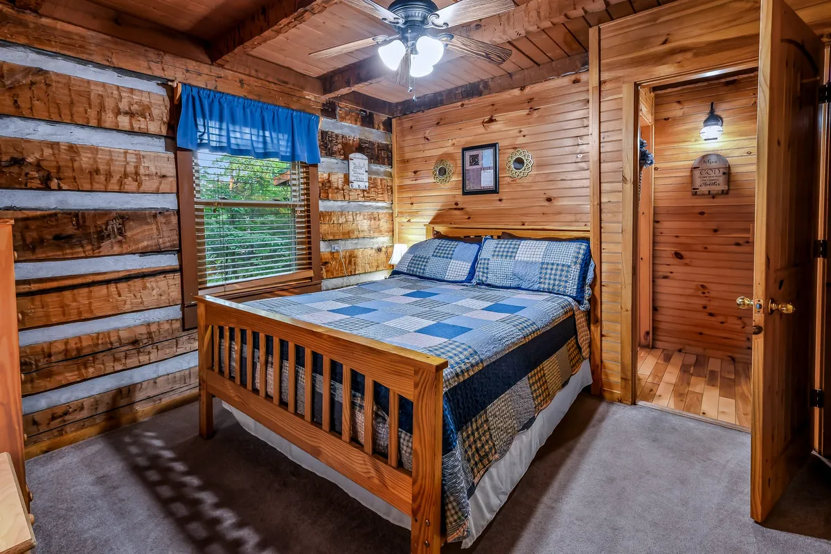 This bedroom scene showcases a wooden bed with plaid bedding, rustic wooden walls, and a window with blue curtains letting in natural light. The setting feels warm and inviting, with decorative wall hangings adding character.