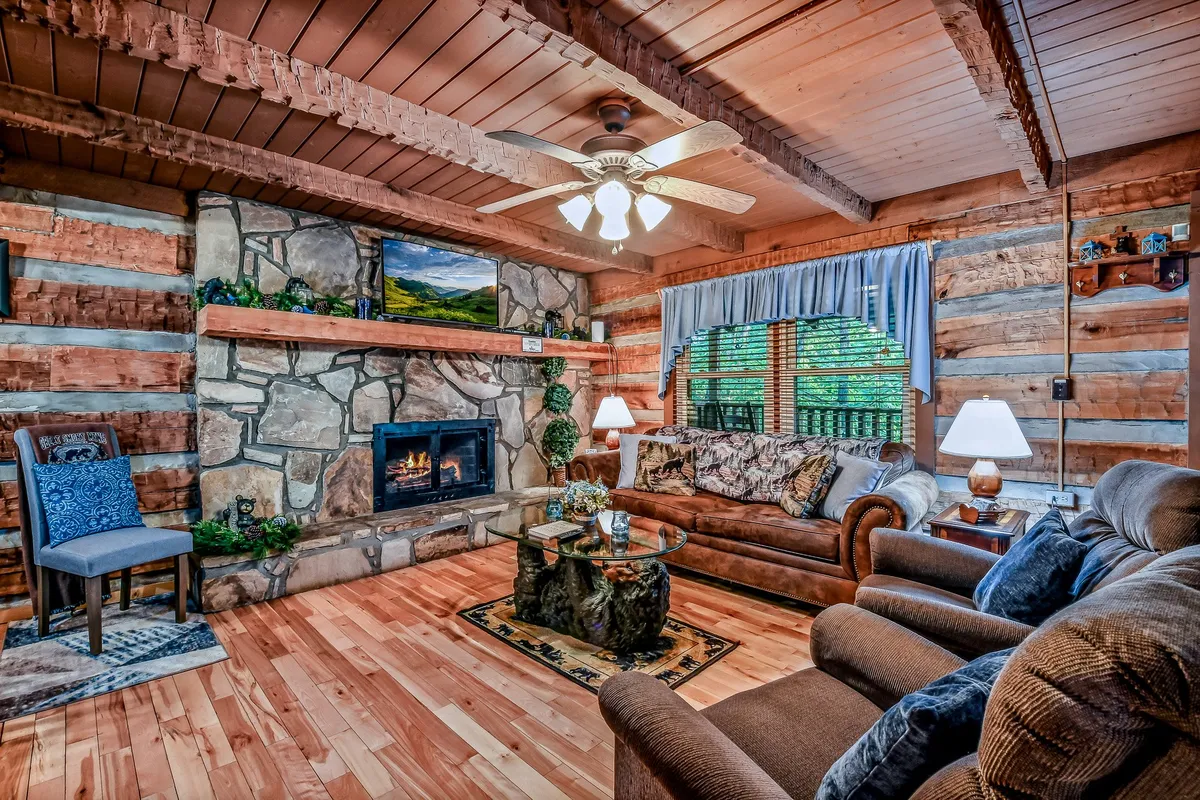 A warmly lit living room featuring a striking stone fireplace, comfortable seating, a wooden coffee table, and exposed wood beams. The space exudes an inviting atmosphere perfect for relaxation, reading, or socializing around the glow of the fire.